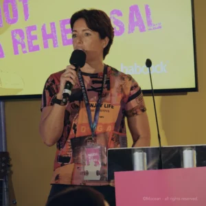 Corporate events london. Company culture. A speaker holding a mic on stage in front of a lectern and with event theming in the background at a punk-themed corporate conference.